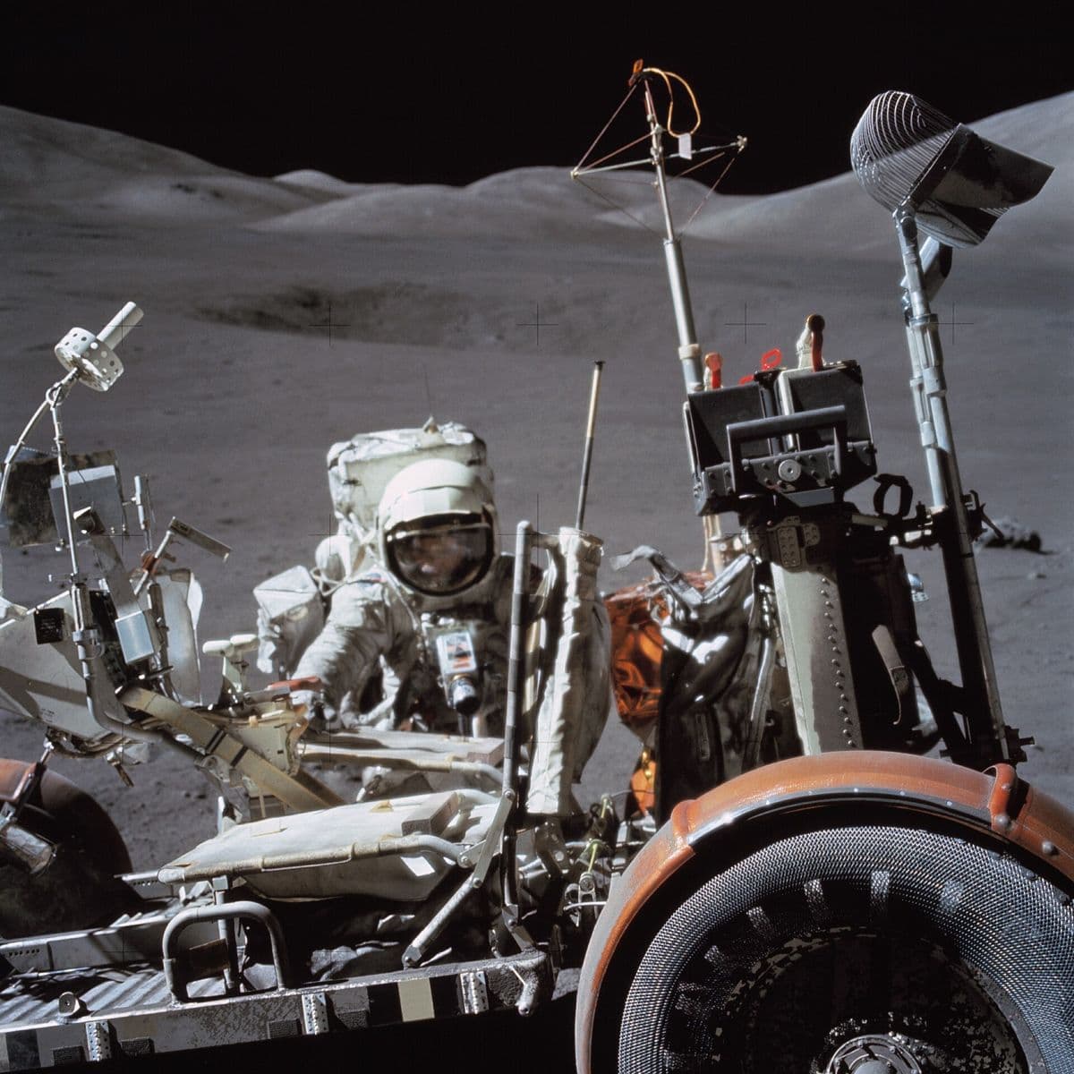 Close view of an Apollo 17 astronaut beside the lunar rover, its wire-mesh wheel and orange fender in the foreground.