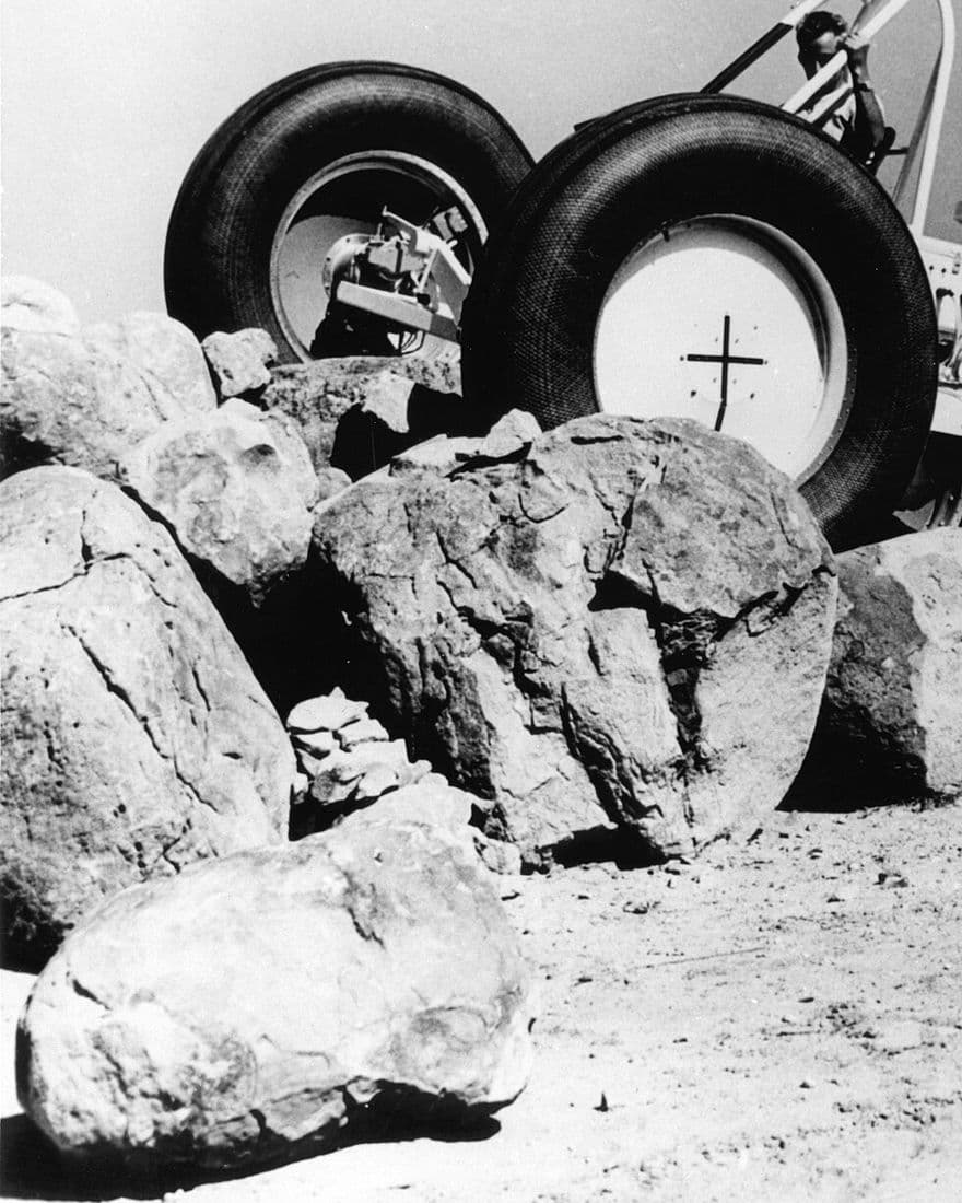 Black-and-white photo of an early rover test vehicle with large tyres climbing over boulders, a driver gripping its roll bar.