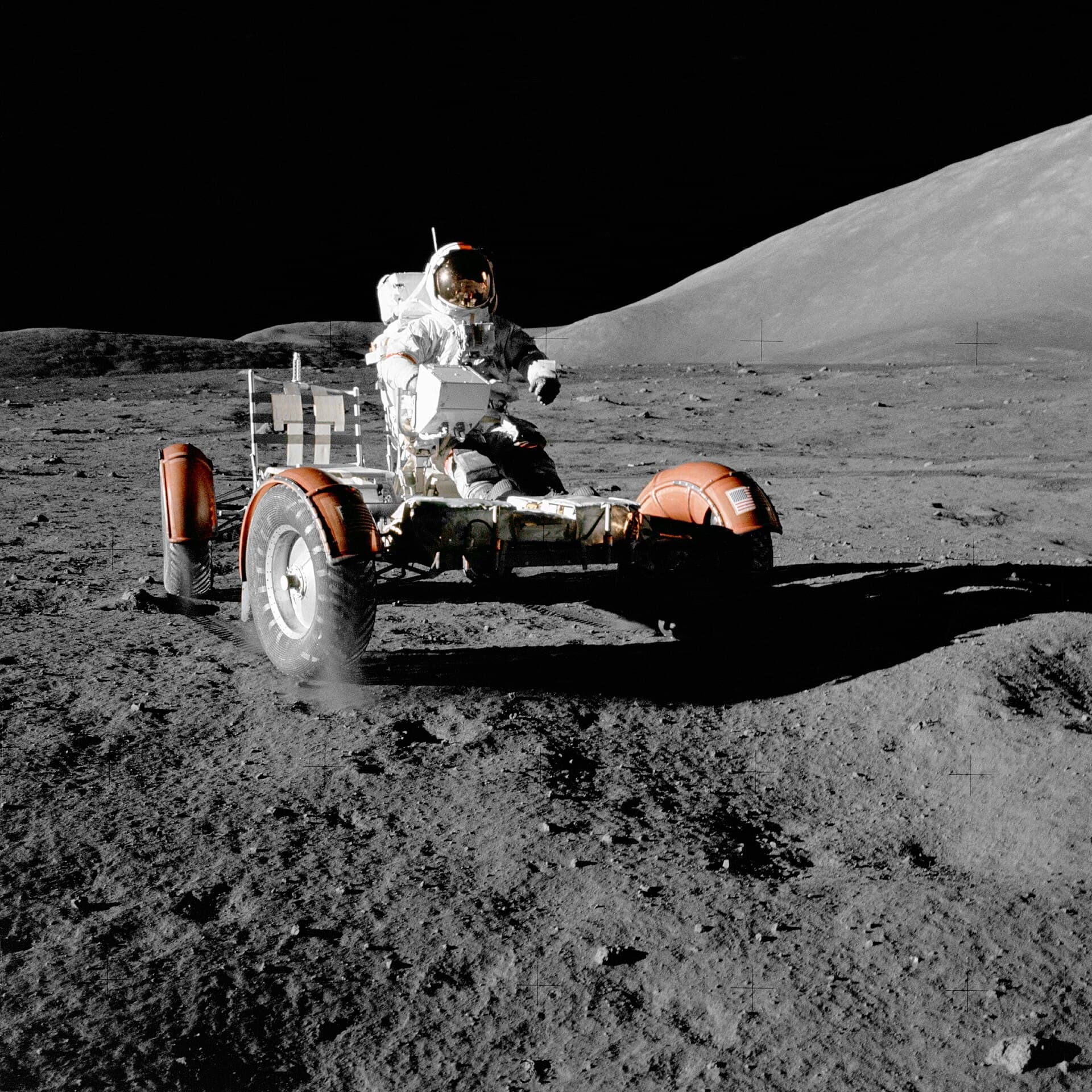 An astronaut in a white spacesuit drives Apollo 17’s Lunar Roving Vehicle, a four-wheeled open frame with orange fenders, across grey lunar soil below a sunlit hill and a black sky.